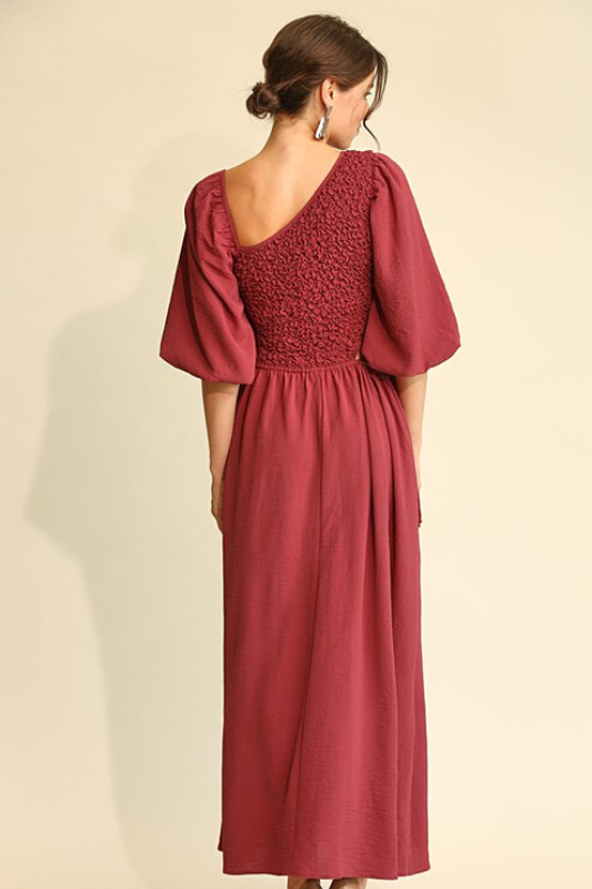 Woman wearing a wine colored midi dress, full aline bottom, smocked middle, asymetric neckline and elbow length balloon sleeves. This is a back view of the item.