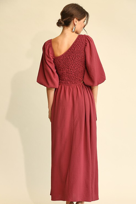 Woman wearing a wine colored midi dress, full aline bottom, smocked middle, asymetric neckline and elbow length balloon sleeves. This is a back view of the item. 
