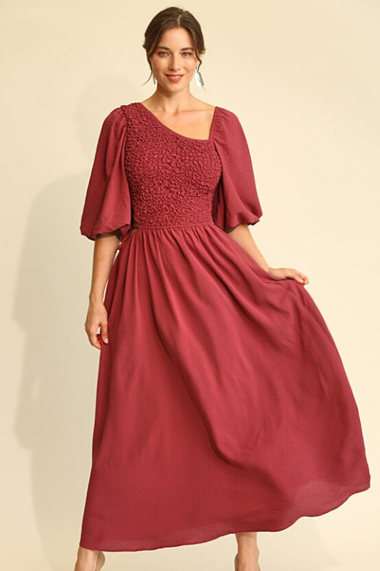 Woman wearing a wine colored midi dress, full aline bottom, smocked middle, asymetric neckline and elbow length balloon sleeves.